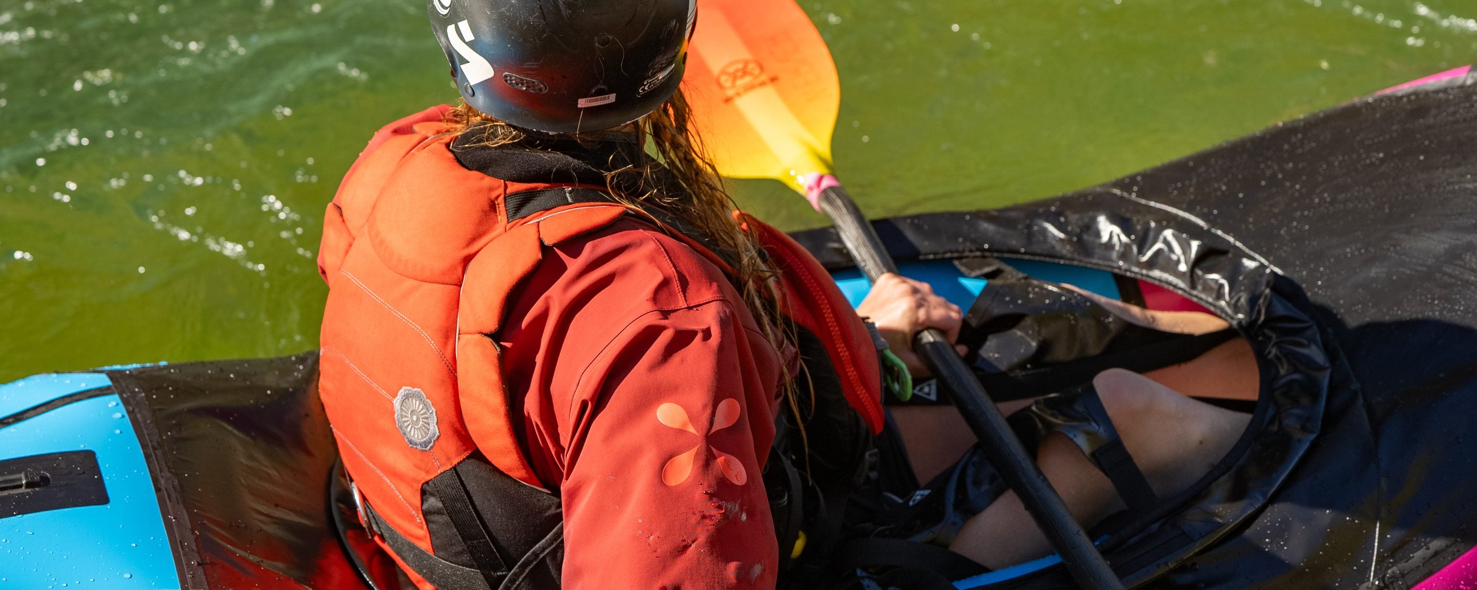An overhead view of a whitewater paddler wearing a red Level Six dry top and an orange PFD, seated in a spray skirt on a blue packraft in green water.