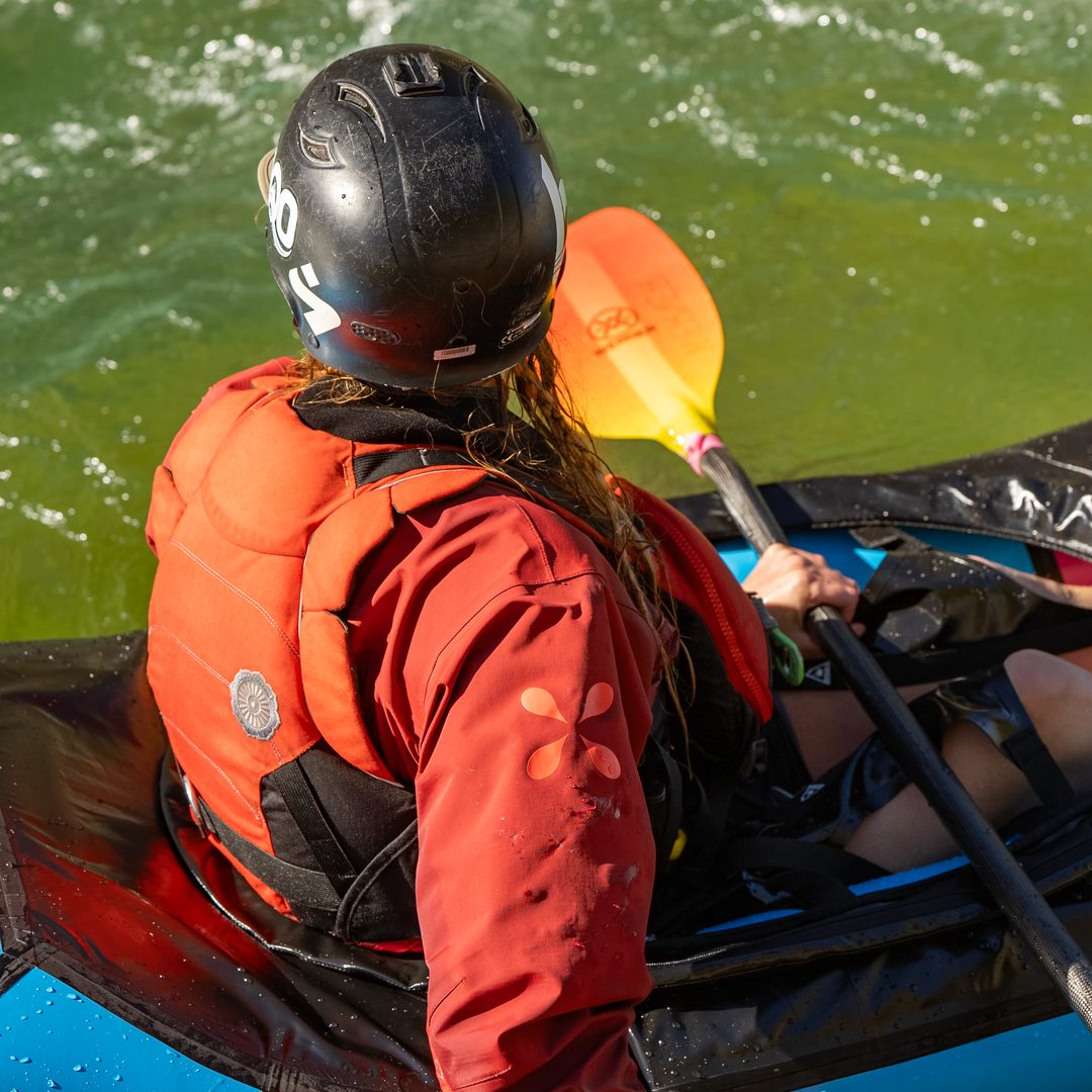 An overhead view of a whitewater paddler wearing a red Level Six dry top and an orange PFD, seated in a spray skirt on a blue packraft in green water.