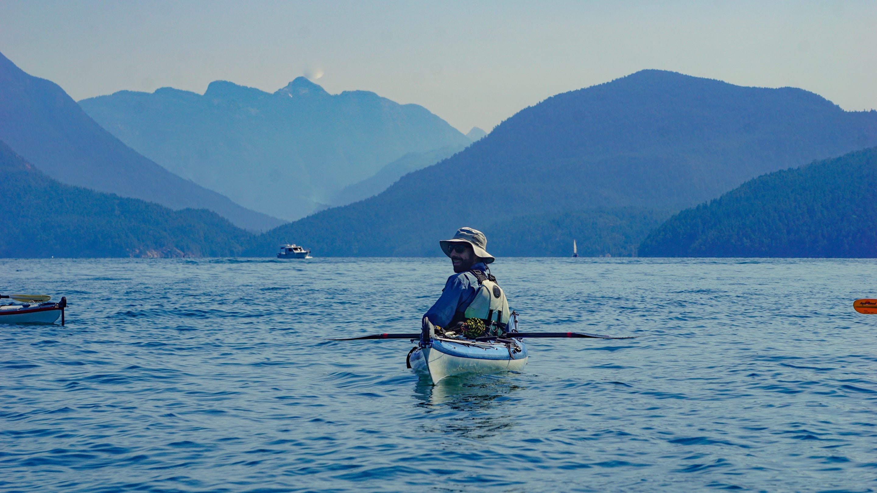 A sea kayaker in a wide-brimmed hat pauses on flat water in Desolation Sound, British Columbia, looking back over one shoulder with forested coastal mountains behind.