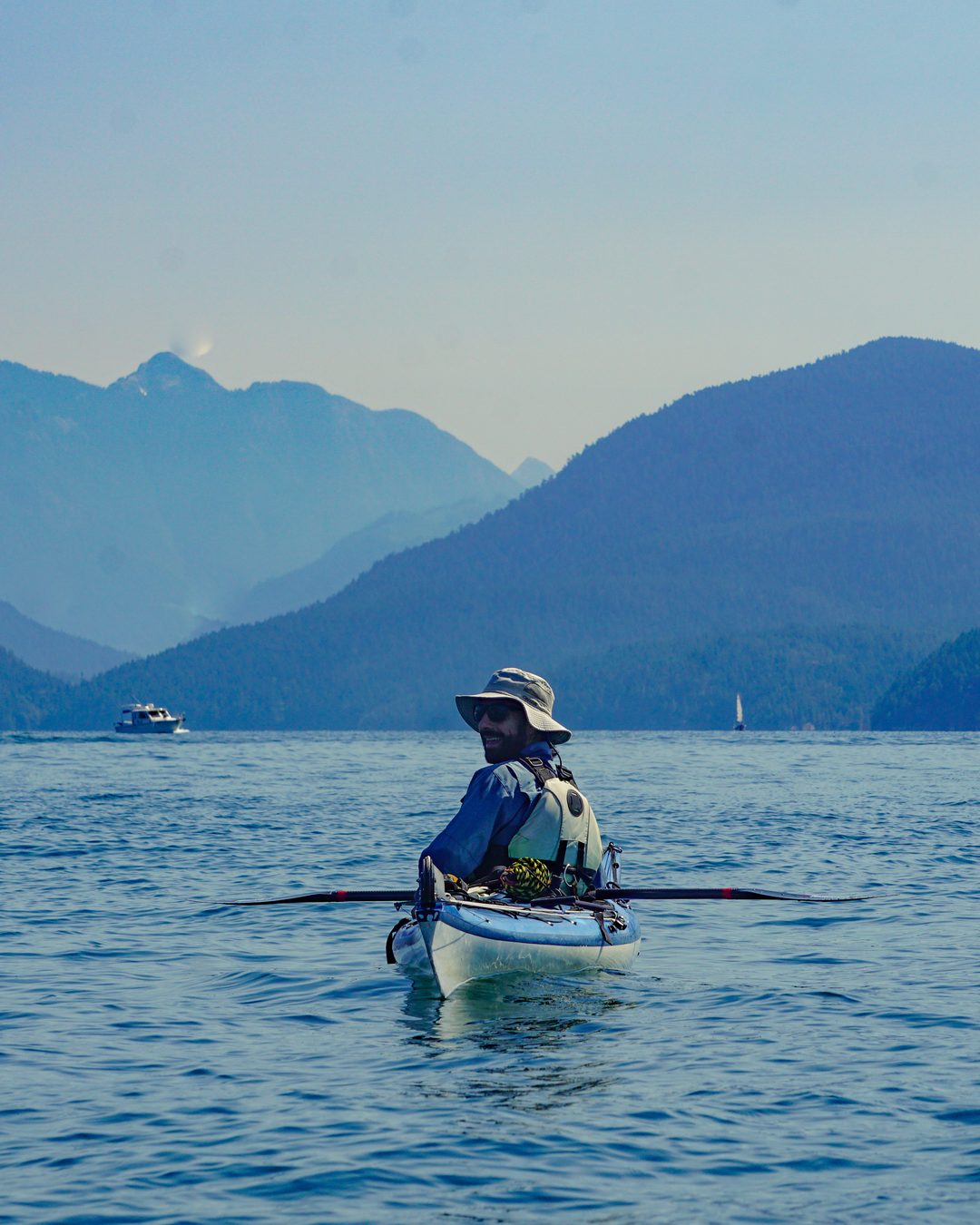 A sea kayaker in a wide-brimmed hat pauses on flat water in Desolation Sound, British Columbia, looking back over one shoulder with forested coastal mountains behind.
